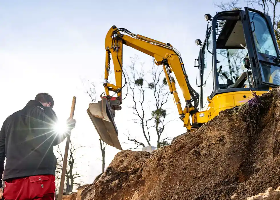 Arbeiter in einem ausgehobenen Erdloch mit Bagger im Hintergrund