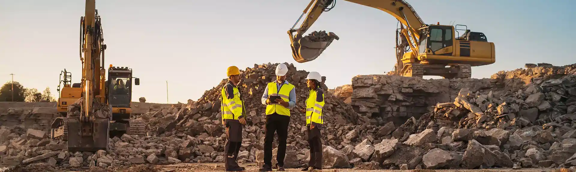 3 BauarbeiterInnen auf einer Baustelle mit 2 Baggern im Hintergrund