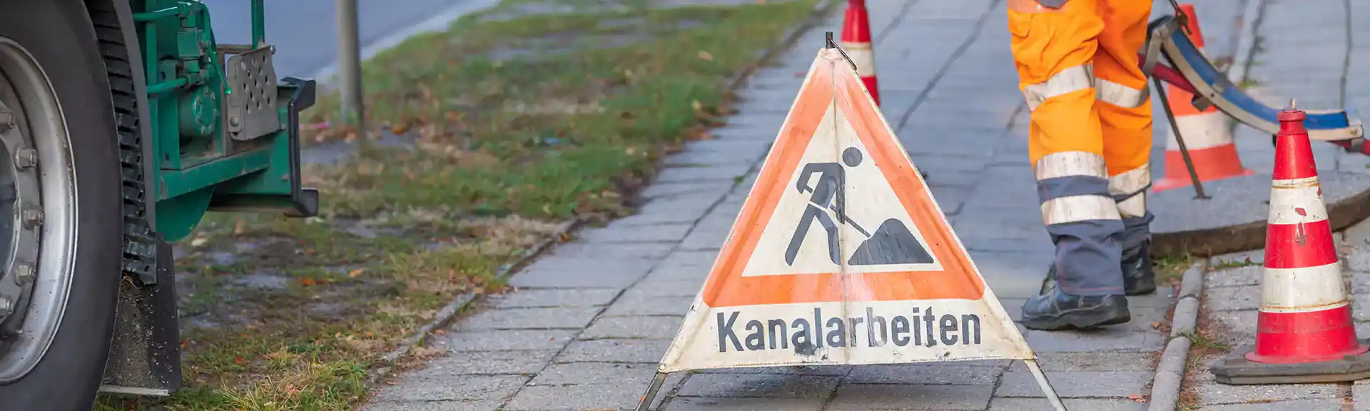 Hinweisschild/Warndreieck Kanalarbeiten. Im Hintergrund Arbeiter in orangener Arbeitskleidung